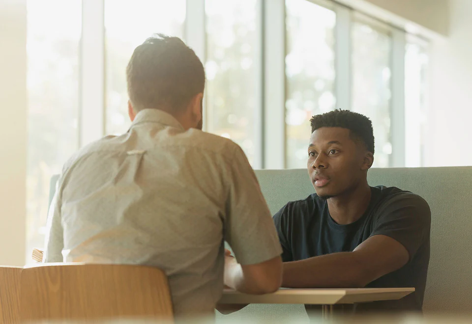 Two men talk across a small table by a sunlit window, one leaning in to listen