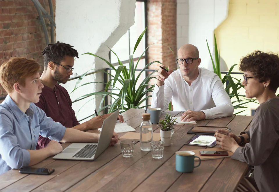 Four colleagues talk over a plan at a long wooden table set with a laptop and coffee mugs