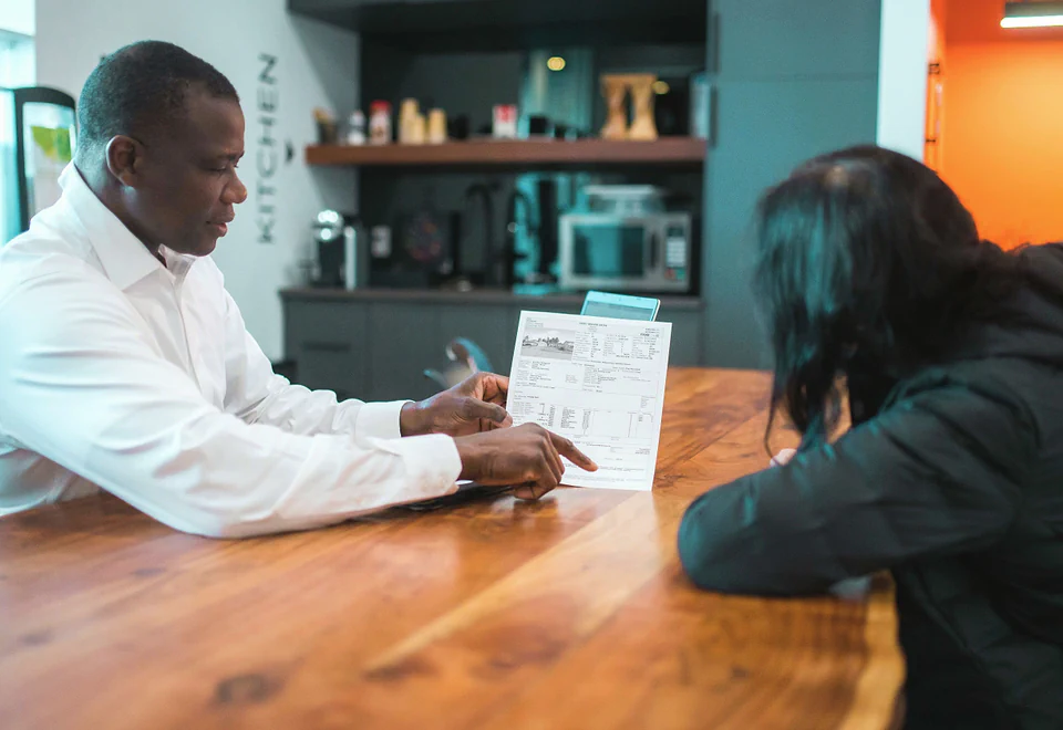 A man points to a line on a printed page as he walks a colleague through it at a wooden table